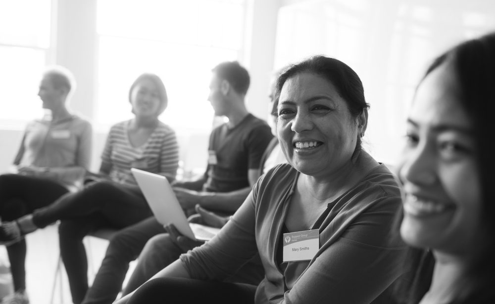 A group of people sits in a circle, engaged in conversation, fostering a sense of community. The focus is on a woman smiling warmly, her name tag a subtle detail. The black-and-white image captures an airy and harmonious space.