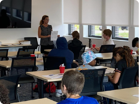 A woman stands at the front of a classroom, holding a document and engaging in group coaching with seated students who attentively take notes. The room is equipped with desks, chairs, and a large screen on the wall, fostering an interactive learning environment.