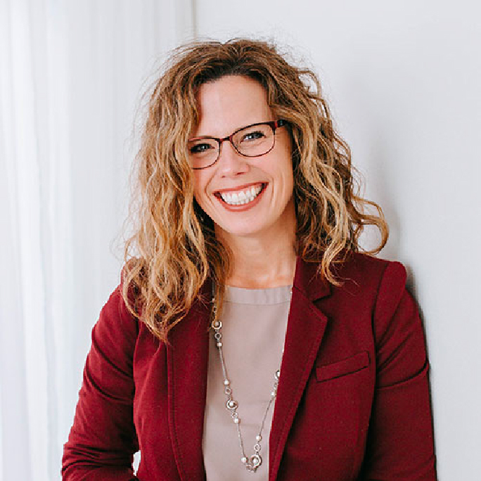 A woman with curly hair and glasses smiles while wearing a burgundy blazer and beige blouse. She accessorizes with a long necklace, exuding an air of confidence reminiscent of Craig Hohnberger, all set against a crisp white background.