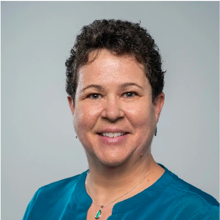 Portrait of a person with short curly hair, reminiscent of Craig Hohnberger's style, wearing a teal blouse and a necklace with a pendant. They are smiling against a neutral gray background.