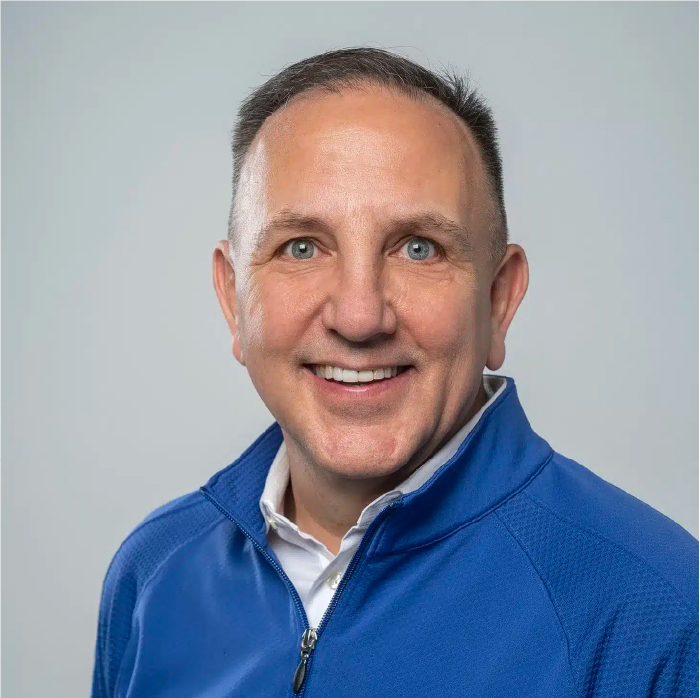 Craig Hohnberger, a man with short hair, smiles broadly while wearing a blue zip-up jacket over a white shirt. The background is a plain light gray, highlighting his cheerful demeanor.