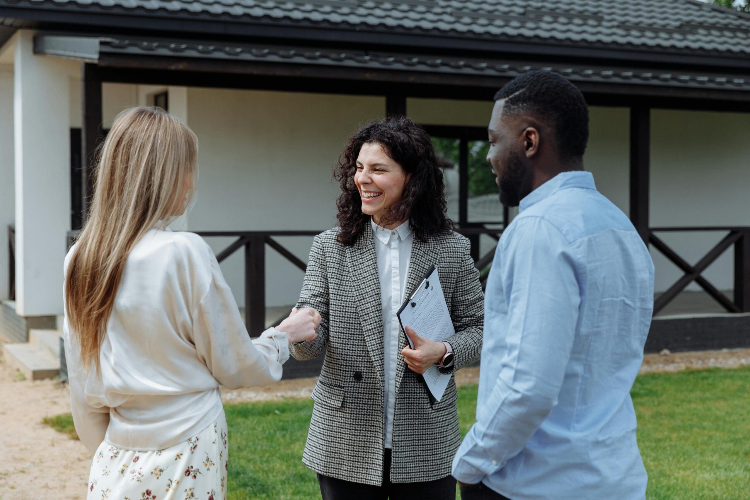 A real estate agent holding documents smiles and shakes hands with a woman as a man stands beside them outside a house with a porch, celebrating the best deal to sell your business or home.